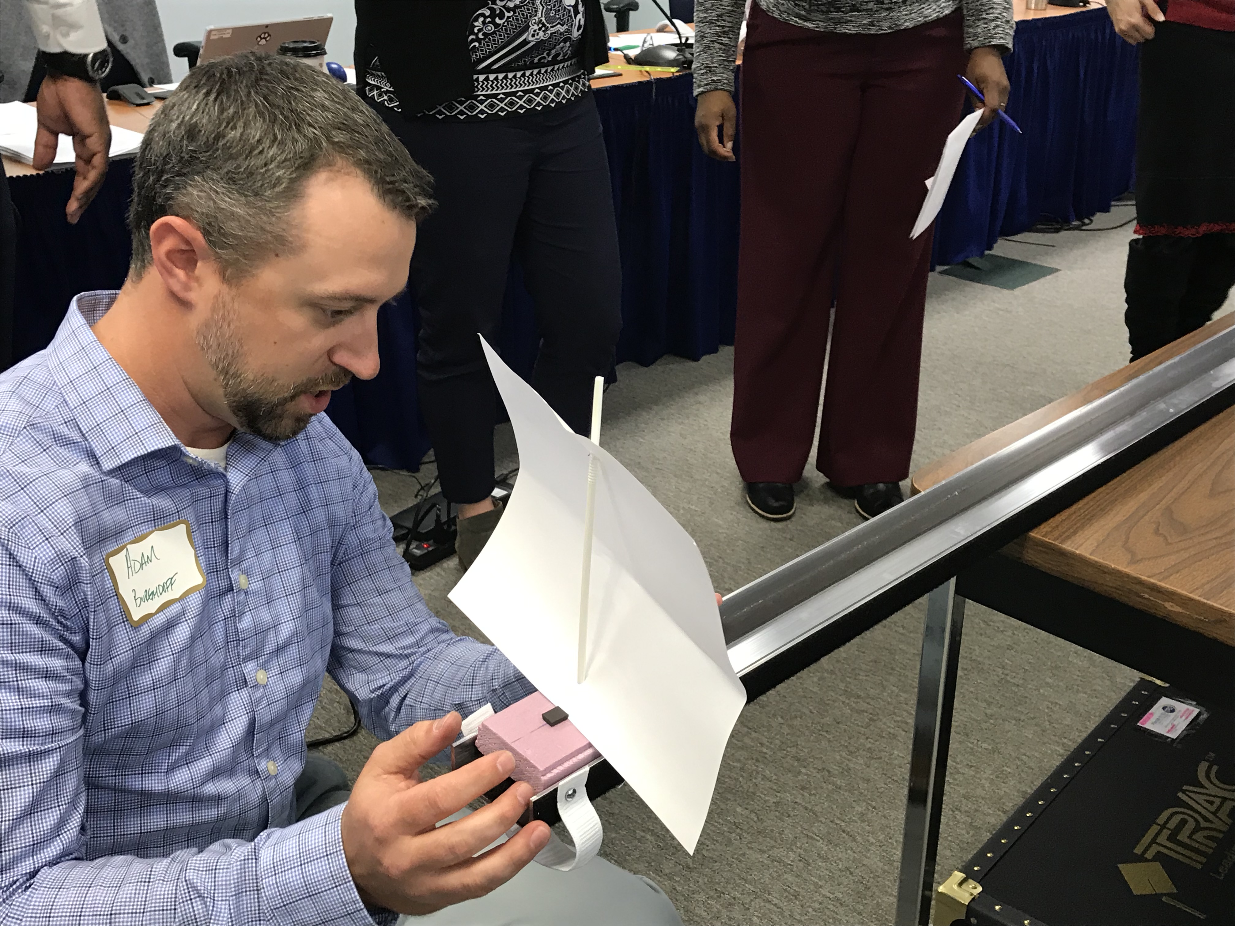 Adam Burghdoff pushing a homemade boat made of a pink styrofoam brick with a paper sail and a wood dowel down a metal track
