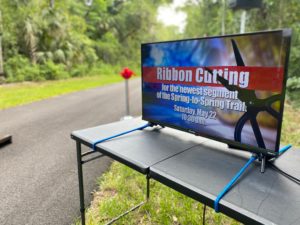TV with words Ribbon cutting for the newest segment of the spring to spring trail saturday May 22 10:30 a.m. and the backdrop is a paved trail surrounded by trees