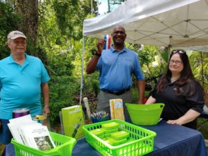 Bob Storke, Stephan Harris and Pamela Blankenship standing behind the River to Sea TPO booth with Stephan holding a piece of the red, white and blue ribbon from the ribbon cutting of the trail